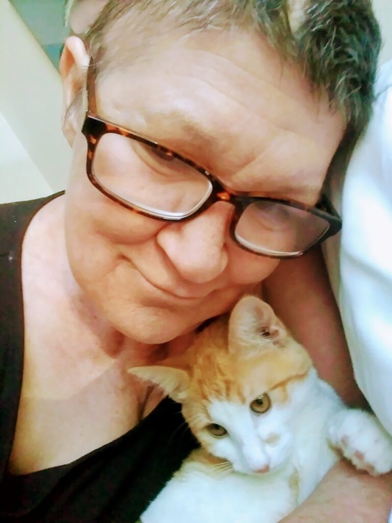 A selfie of a white woman with short brown hair and brown tortoise glasses in a black t-shirt holding a orange and white cat.