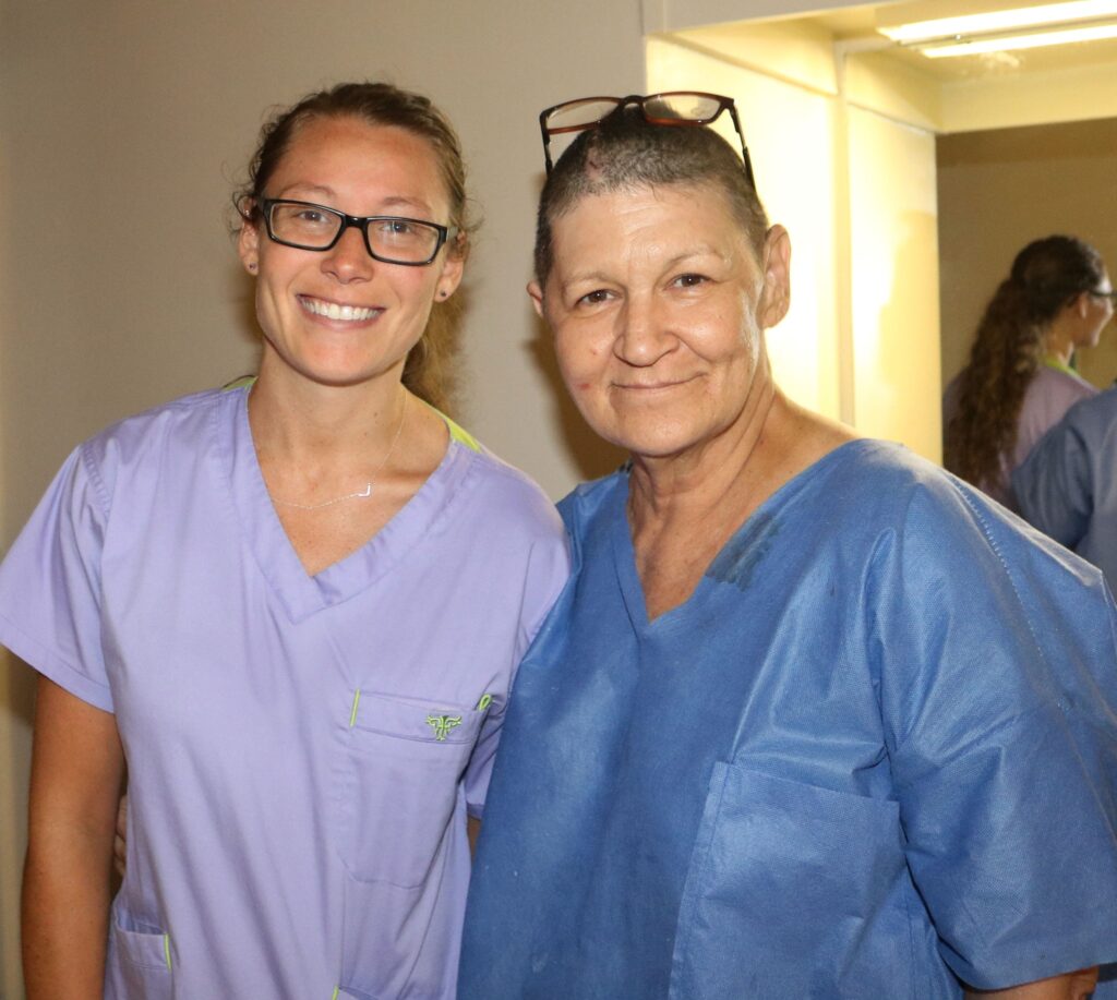A white woman with brown hair and black glasses stands on the left in light purple scrubs. A white woman with a shaved head and glasses on her head in blue scrubs stands on the right. They are standing in front of a light yellow painted hallway.