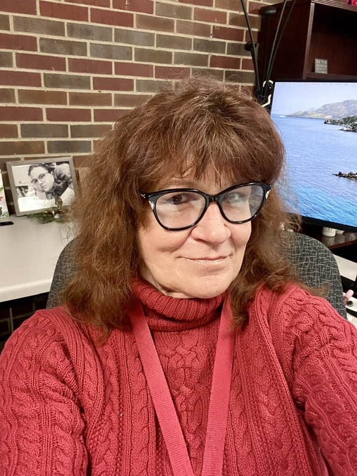 A selfie of a white woman with brown/red hair and black cat eye glasses in a dark pink sweater sitting at her desk with a red brick wall in the background.