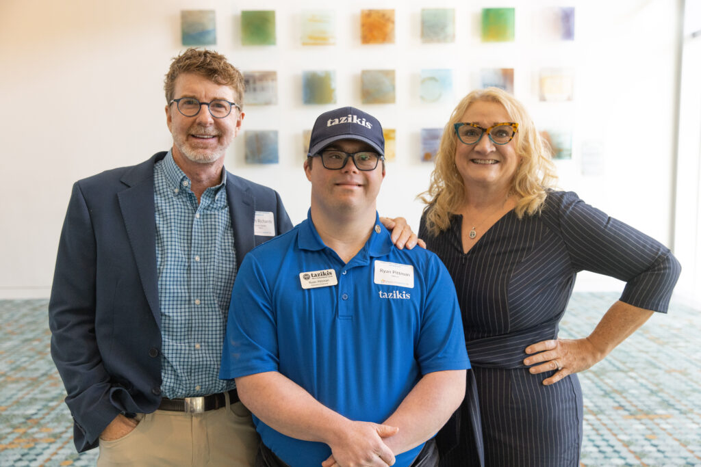 Three individuals pose for a photo indoors in front of a white wall adorned with colorful square art pieces. On the left, a man in a blazer and glasses smiles warmly. In the center, a man wearing a blue Taziki’s shirt and cap stands confidently. On the right, a woman in a striped dress and glasses stands with her hand on her hip, smiling brightly.
