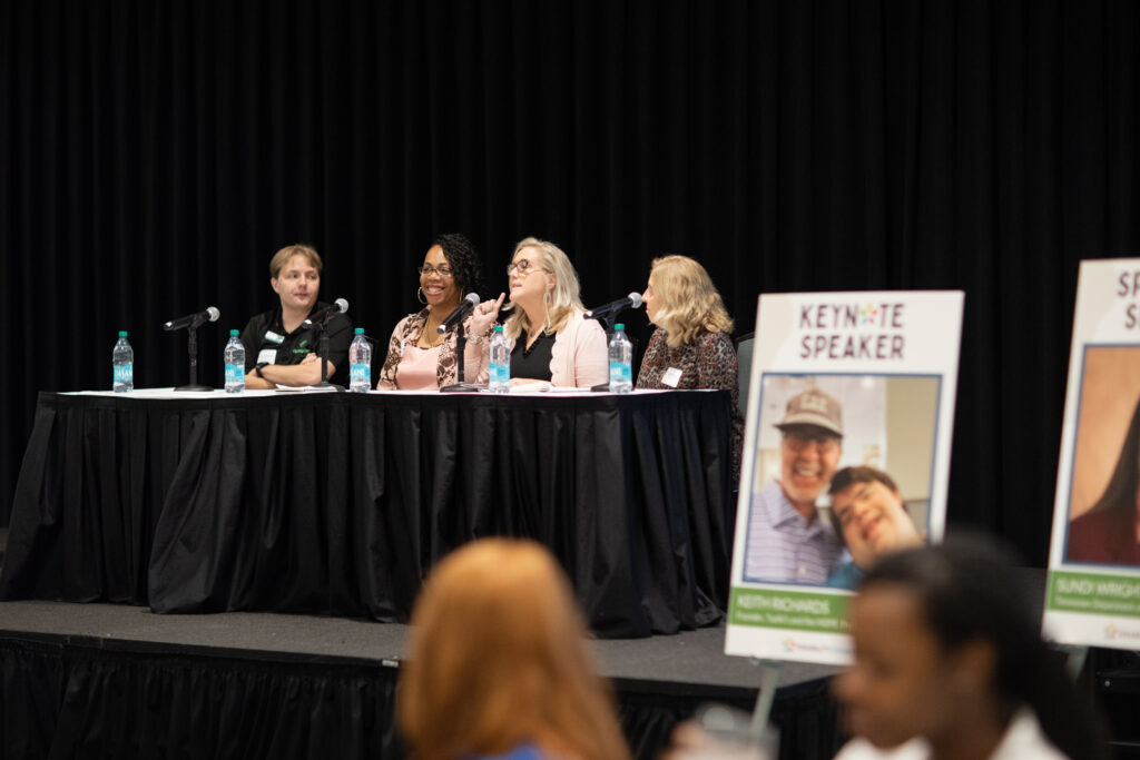 A panel of four people sits at a long table with microphones during a conference. They are engaged in discussion. The backdrop is a dark curtain, and there are posters and an audience visible in the foreground.